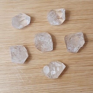 Peut inclure: Six cristaux de quartz transparents, chacun avec une forme et une taille uniques, sont disposés sur une surface en bois brun clair. Les cristaux sont translucides et ont un léger éclat.