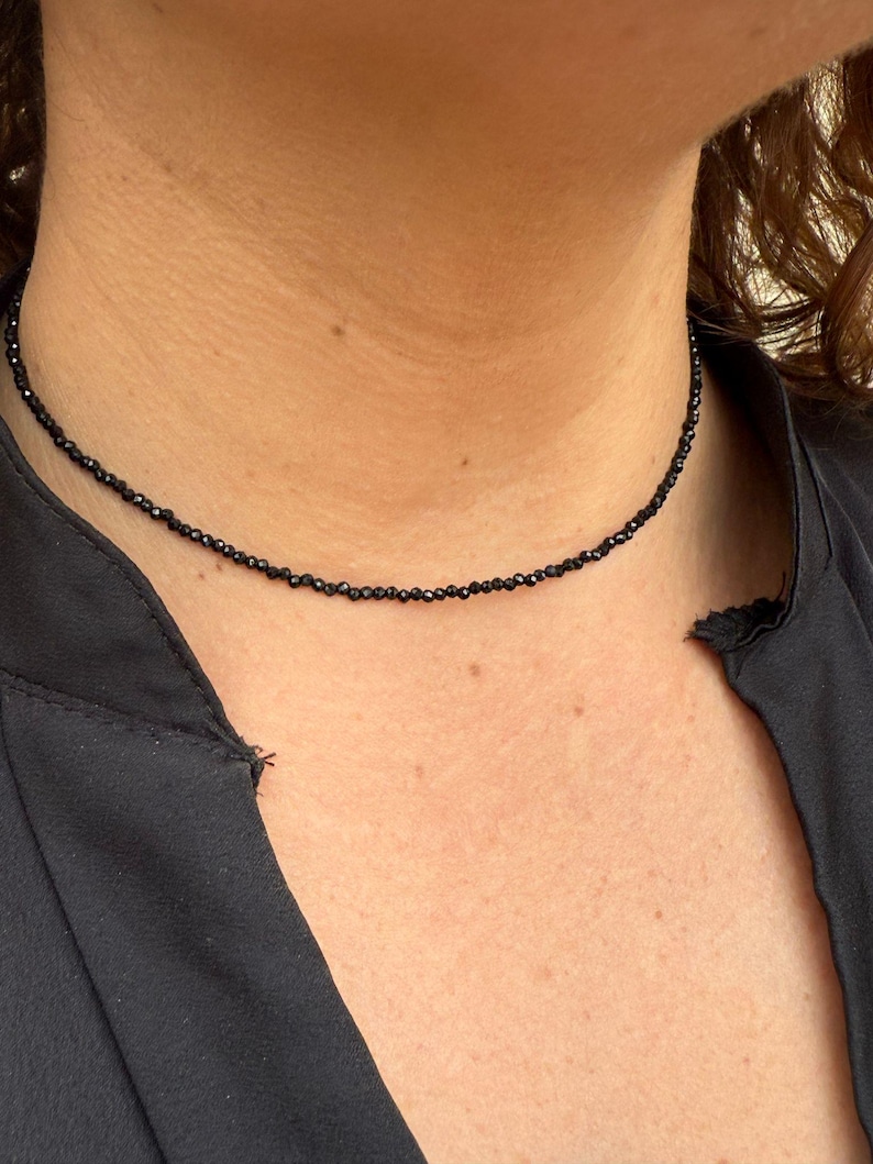 May include: A fine black beaded choker necklace. The necklace features small, dark beads, strung together to form a simple, elegant design. The necklace sits close to the neck, offering a touch of sophistication.