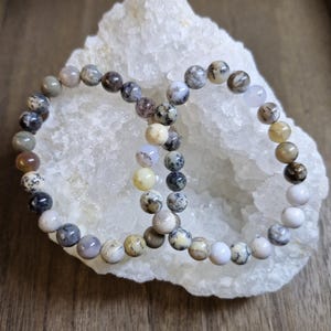 Peut inclure: Deux bracelets de perles aux couleurs variées, dont blanc, gris, marron et jaune, sont présentés sur une surface cristalline blanche. Les bracelets, composés de petites perles rondes, reposent sur un fond en bois.