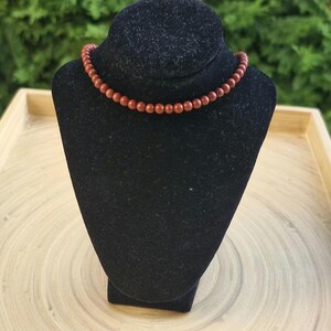 May include: A necklace with small, round, reddish-brown beads. The beads are strung together to form a choker-style necklace. The necklace is displayed on a black velvet jewelry bust against a blurred green background.