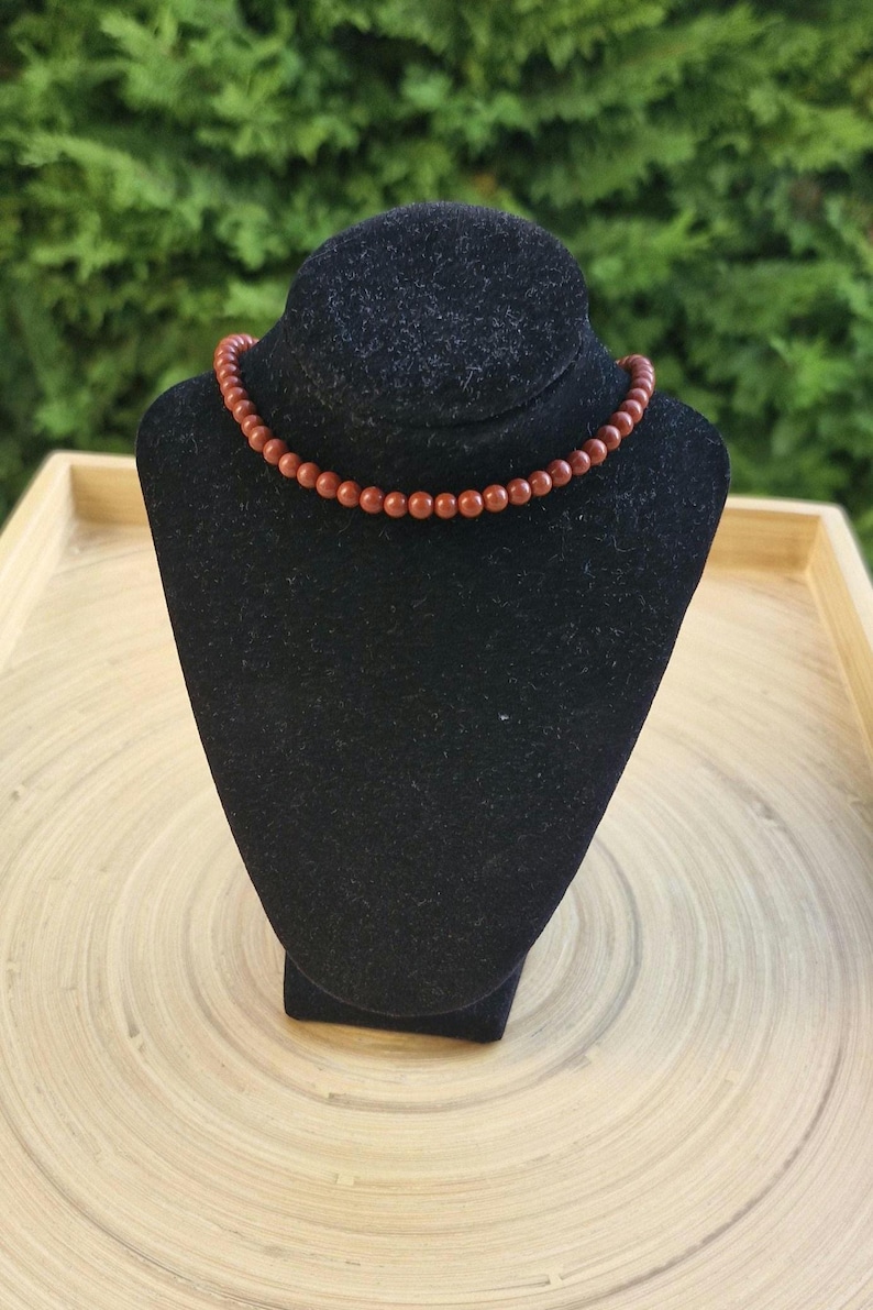 May include: A necklace with small, round, reddish-brown beads. The beads are strung together to form a choker-style necklace. The necklace is displayed on a black velvet jewelry bust against a blurred green background.