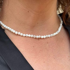 Peut inclure: Un collier ras du cou composé de petites perles blanches rondes. Les perles sont enfilées ensemble, créant une chaîne continue. Le collier est porté autour du cou. La personne porte un haut noir. Les perles semblent être de taille et de forme uniformes.