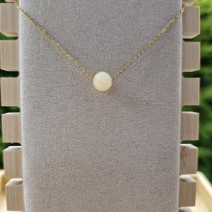 Op de afbeelding: Een delicate goudkleurige ketting met een enkele, ronde, cr&egrave;mekleurige kralen hanger. De ketting wordt gepresenteerd tegen een neutrale achtergrond, met een houten displaystandaard aan de linkerkant. De ketting is fijn en elegant.