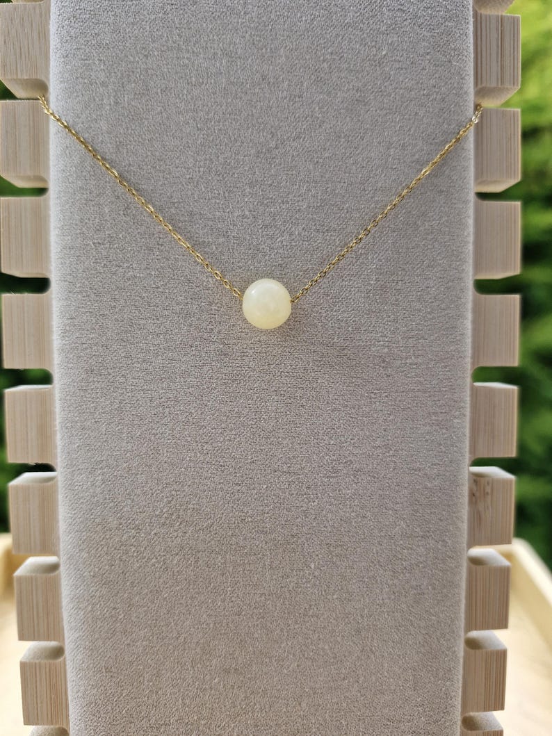 Op de afbeelding: Een delicate goudkleurige ketting met een enkele, ronde, cr&egrave;mekleurige kralen hanger. De ketting wordt gepresenteerd tegen een neutrale achtergrond, met een houten displaystandaard aan de linkerkant. De ketting is fijn en elegant.