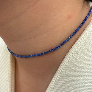 May include: A delicate necklace composed of small, faceted blue beads. The necklace is worn around the neck, resting against a cream-colored sweater. The beads appear to be evenly sized and spaced, creating a subtle, elegant accessory.