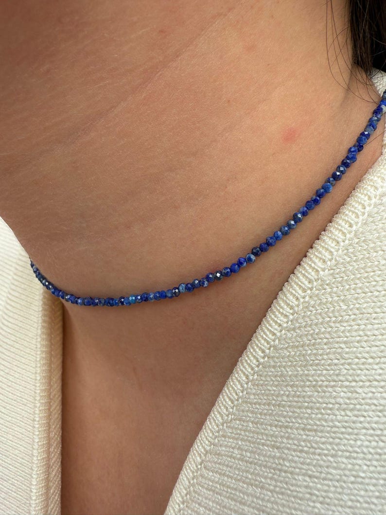 May include: A delicate necklace composed of small, faceted blue beads. The necklace is worn around the neck, resting against a cream-colored sweater. The beads appear to be evenly sized and spaced, creating a subtle, elegant accessory.