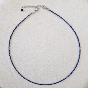 May include: A delicate necklace featuring small, dark blue beads strung together. The necklace has a silver-colored clasp and an adjustable chain. The beads are evenly spaced, creating a simple yet elegant design. The necklace is displayed on a white surface.