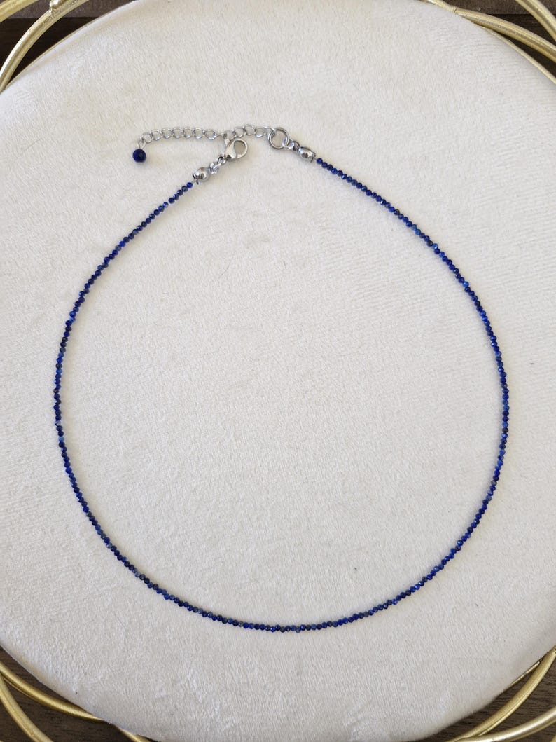 May include: A delicate necklace featuring small, dark blue beads strung together. The necklace has a silver-colored clasp and an adjustable chain. The beads are evenly spaced, creating a simple yet elegant design. The necklace is displayed on a white surface.