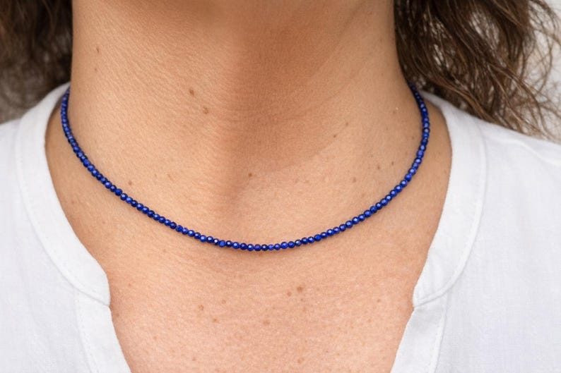 May include: A delicate, dark blue beaded necklace. The necklace is composed of small, round beads, creating a simple yet elegant design. The necklace is worn around the neck, against a white shirt.