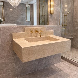 May include: A modern bathroom featuring a beige stone sink with gold fixtures. The sink is mounted on a wall with gray marble tiles. A large mirror and a chandelier are visible in the background.