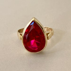May include: A gold ring featuring a large, teardrop-shaped red gemstone.