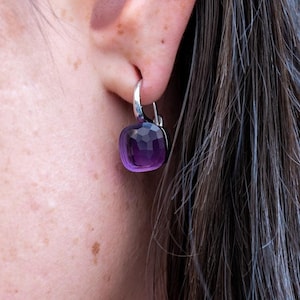 May include: A close-up of a woman's ear with a silver hoop earring with a purple gemstone dangling from it.