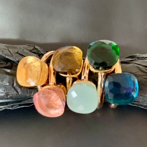 May include: A set of six gold rings with colourful gemstone settings. The rings feature a variety of colours, including green, blue, yellow, and pink.