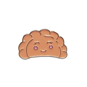 May include: A light brown enamel pin shaped like a folded dumpling or pierogi. The design features a simple, smiling face with closed eyes, curved eyelashes, and small pink circular cheeks, all outlined in a thin, dark metallic border.