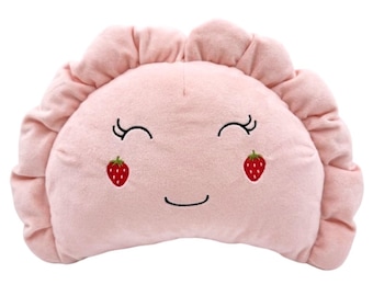 May include: A semi-circular plush pillow in a soft pastel pink color featuring a ruffled edge. The front displays an embroidered face with closed eyes, a simple curved mouth, and two small red strawberry icons positioned as rosy cheeks.