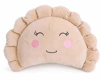 May include: A beige, semi-circular plush pillow shaped like a dumpling. The pillow features embroidered closed eyes with eyelashes, a simple curved mouth, and two round pink cheeks stitched onto the center of the soft, textured fabric surface.