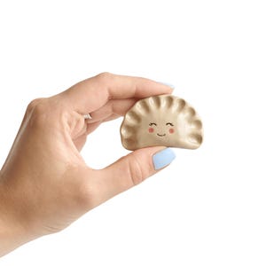 May include: A tan dumpling with a smiling face, held against a white background. The dumpling has a crimped edge and painted details for the eyes and mouth. The image is of a food-themed item.