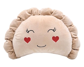 May include: A beige plush pillow shaped like a dumpling with a ruffled edge. The front features an embroidered face with closed eyes, a small curved smile, and two red heart-shaped cheeks. The soft fabric has a smooth texture throughout.