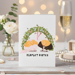 May include: A white greeting card with a wedding-themed illustration. The card features two cartoon figures under a floral arch, with the text "PERFECT MATCH" below. A glass of champagne and a bouquet of white roses are in the background.