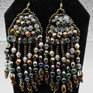 Puede incluir: Un par de pendientes colgantes con forma de lágrima. Los pendientes están hechos de cuentas plateadas, doradas y negras. Las cuentas están dispuestas en forma de cascada.
