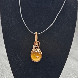 Può includere: Una collana con un ciondolo avvolto in filo dorato e una pietra rotonda di occhio di tigre gialla. La collana è su un cordoncino nero.
