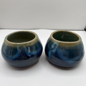May include: Two small, rounded ceramic bowls featuring a glossy glaze that transitions from a deep navy blue at the base to a lighter, muted sage green around the circular rim. The bowls have a smooth, curved silhouette and a wide, open top.