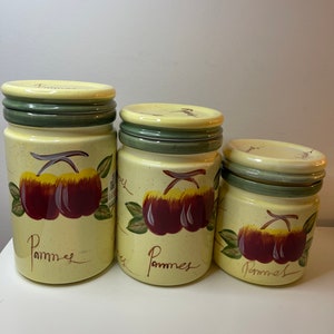 May include: A trio of ceramic kitchen storage jars. The jars are pale yellow with green rims and lids. Each jar has a painted design of red apples with green leaves and the word "Pommes" in script.