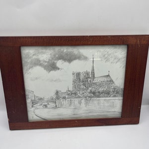 May include: A framed pencil sketch of Notre Dame Cathedral in Paris. The artwork depicts the cathedral's facade, the Seine River, and a bridge. The frame is made of dark brown wood.
