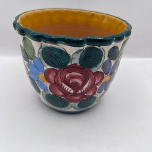 Puede incluir: Una maceta de cerámica con borde festoneado. La maceta está decorada con una gran rosa burdeos, flores azules y moradas y hojas verdes sobre un fondo blanco. El borde es amarillo y el borde es verde.