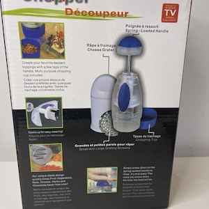 May include: A product box for a kitchen chopper and cheese grater. The box features images of the product and its components, including a spring-loaded handle and chopping cup. Text on the box describes the product's features and ease of use.