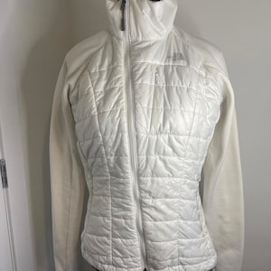 Puede incluir: Chaqueta acolchada blanca con cuello alto y cremallera completa. La chaqueta tiene un material tipo forro polar en las mangas y los laterales. El logotipo de The North Face es visible en la parte superior del pecho.