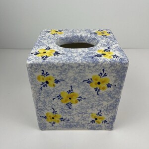 May include: Square ceramic tissue box. It features a light blue speckled background and a yellow floral pattern. The tissue box has a square opening on top. A decorative item for the bathroom or bedroom.