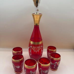 Puede incluir: Un juego de decantador de vidrio rojo con tapón y adornos dorados. El decantador y seis vasos a juego presentan diseños florales plateados y dorados ornamentados. El juego está diseñado para servir bebidas.