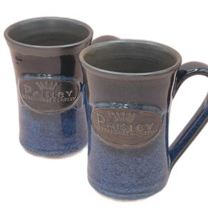 Może przedstawiać: Dwa niebieskie ceramiczne kubki o teksturowanym wykończeniu. Każdy kubek ma ciemnoniebieską obręcz i uchwyt, z jaśniejszą niebieską podstawą. Kubki mają brązowy owal z napisem "Paisley Coffeehouse Eatery".