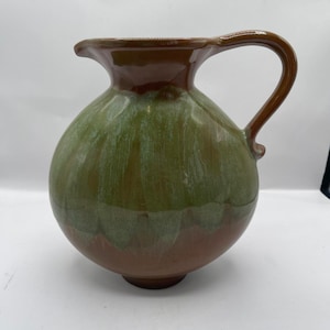 Puede incluir: Jarra de cerámica hecha a mano con cuerpo redondeado y asa curva. La jarra presenta un esmalte de dos tonos, con una mitad inferior verde y una mitad superior y asa marrón. Adecuada para servir bebidas, mide unos 25 cm de altura.