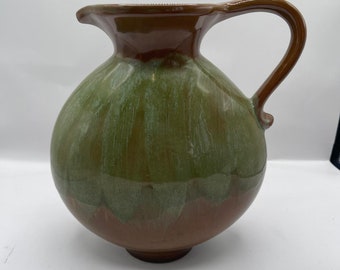 May include: A handcrafted ceramic pitcher with a rounded body and a curved handle. The pitcher features a two-tone glaze, with a green lower half and a brown upper half and handle. The pitcher is suitable for serving beverages.