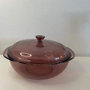 Puede incluir: Una fuente redonda de vidrio transparente con tinte púrpura y tapa a juego. La fuente tiene una base redondeada y un borde ligeramente ensanchado. La tapa presenta un pequeño asa redondeada. La fuente está diseñada para cocinar y servir.