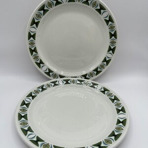 May include: Two white ceramic plates with a decorative border. The border features a repeating pattern of green and blue leaf-like shapes within a geometric design. The plates are round and have a simple, elegant design.