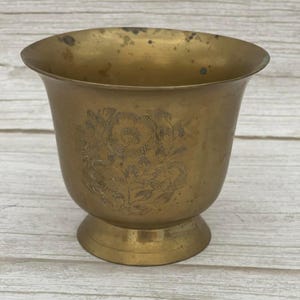 May include: A vintage brass planter with a flared rim and a pedestal base. The planter features an etched floral design on the body. The brass has a warm, golden hue and shows signs of age.