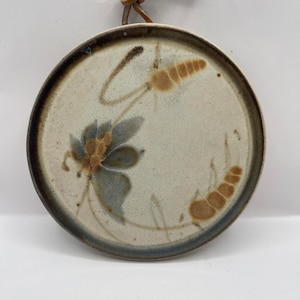 May include: Round ceramic wall hanging with a floral design. The plate has a cream-coloured background with a dark brown rim and a brown leather loop for hanging. The floral design features blue and brown accents.
