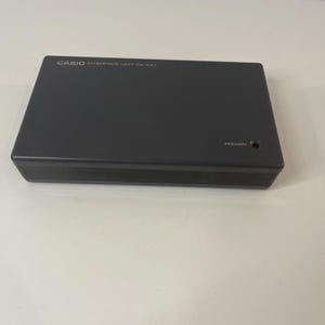 May include: A dark grey Casio Interface Unit PA-100. The rectangular device has the word "POWER" printed on the right side, with a small red light. The top of the device has the words "CASIO INTERFACE UNIT PA-100" printed in white.
