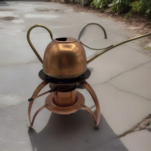 May include: A copper watering can with a long spout and a curved handle. The can sits on a copper and black stand with three curved legs. The spout and handle are brass-coloured.