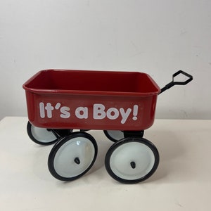 May include: A small, red metal wagon with black wheels and white wheel covers. The wagon has the text "It's a Boy!" in white lettering on the side. The wagon has a handle for pulling.