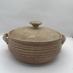 Può includere: Una pirofila in ceramica beige con coperchio. Il piatto ha due manici e un design strutturato e scanalato sui lati. Il coperchio ha un pomello arrotondato. Il piatto viene probabilmente utilizzato per cucinare e servire il cibo.