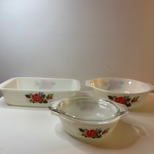 May include: Three vintage white glass baking dishes with a floral rose design. The set includes a rectangular casserole dish, a round bowl, and a small bowl with a glass lid. The roses are red, pink, and blue.
