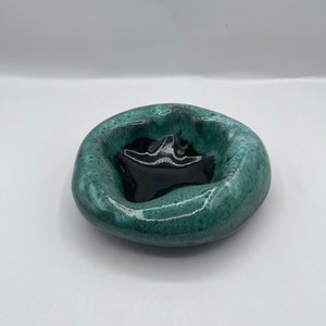 May include: A handmade ceramic ashtray. It has a glossy teal exterior and a dark interior. The ashtray has a rounded, organic shape, with a smooth, glazed finish. The interior features a star-shaped design.