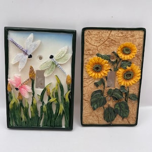 Puede incluir: Dos cubiertas decorativas para interruptores de luz. Una presenta tres libélulas en blanco, rosa y verde sobre un campo de hierba. La otra tiene tres girasoles amarillos con hojas verdes. Ambas tienen un borde verde oscuro.