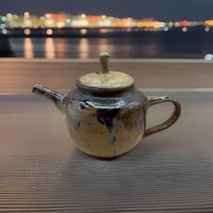 May include: A small, handcrafted ceramic teapot with a lid. The teapot has a rounded body, a curved spout, and a handle. The colors are primarily earthy tones of brown, tan, and a touch of blue. The lid has a small knob on top.