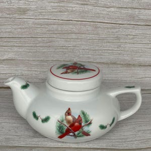 May include: White ceramic teapot with a lid, featuring a red cardinal bird design on the front and lid. The teapot has a curved spout, a handle, and green pine sprig accents. A charming piece for tea lovers.