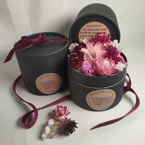 May include: Two black round boxes with lids, each containing dried flowers. The boxes have a brown label with the text "Fragrance & Flowers" and a small illustration of a brick wall with a gate. The box on the left has a burgundy velvet ribbon tied around it. The box on the right has a label with the text "Rose Geranium, Oil Encapsulating Flowers with Artisan Fragrance, Hand Tossed"